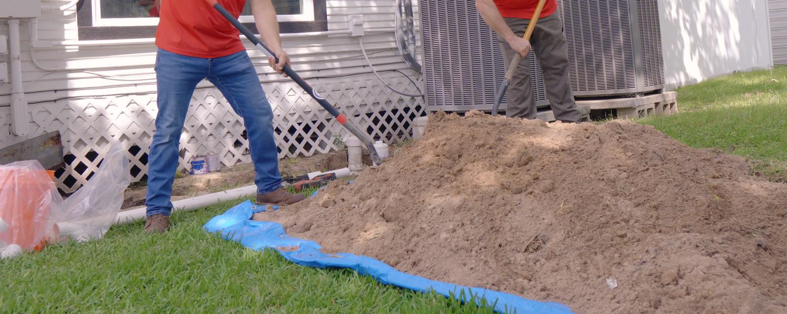 Two plumbers shoveling dirt from an access pit beside a house for a sewer line repair